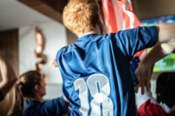 Young man (facing away from the camera) cheers on a team while wearing a player's jersey 
