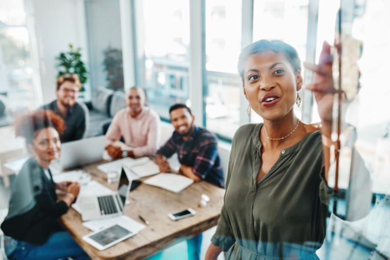 Woman gives a presentation in an office conference room. She is speaking to a table of coworkers.